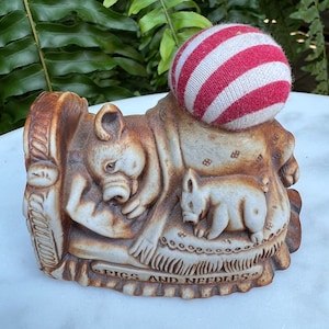 May include: A ceramic figurine of two pigs sleeping in a bed with a red and white striped pincushion on top. The base of the figurine reads "Pigs and Needles".