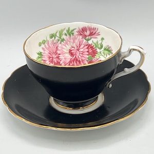 May include: A black teacup and saucer set with a gold trim. The inside of the teacup features a floral design with pink and red flowers and green leaves. The saucer has a scalloped edge.
