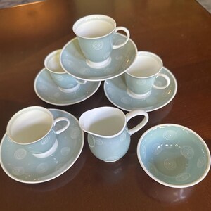 Puede incluir: Un juego de cuatro tazas de té de cerámica azul claro con sus platos, una jarra para crema y un cuenco pequeño. Cada pieza presenta un patrón de espirales blancas. Las tazas tienen asas curvas e interiores blancos, con bordes a juego en todo el set.