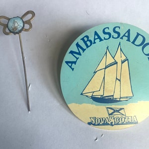 May include: A blue and white pin with a sailboat and the text "Ambassador Nova Scotia".