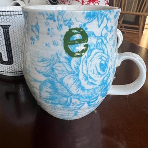 K&ouml;nnte beinhalten: Wei&szlig;er Keramikbecher mit blauem Blumenmuster und einem gr&uuml;nen Buchstaben "e". Der Becher hat einen gro&szlig;en Henkel und geh&ouml;rt zu einer Sammlung von Bechern mit verschiedenen Mustern und Farben. Andere Becher im Hintergrund haben rote Blumenmuster und schwarz-wei&szlig;e geometrische Designs.
