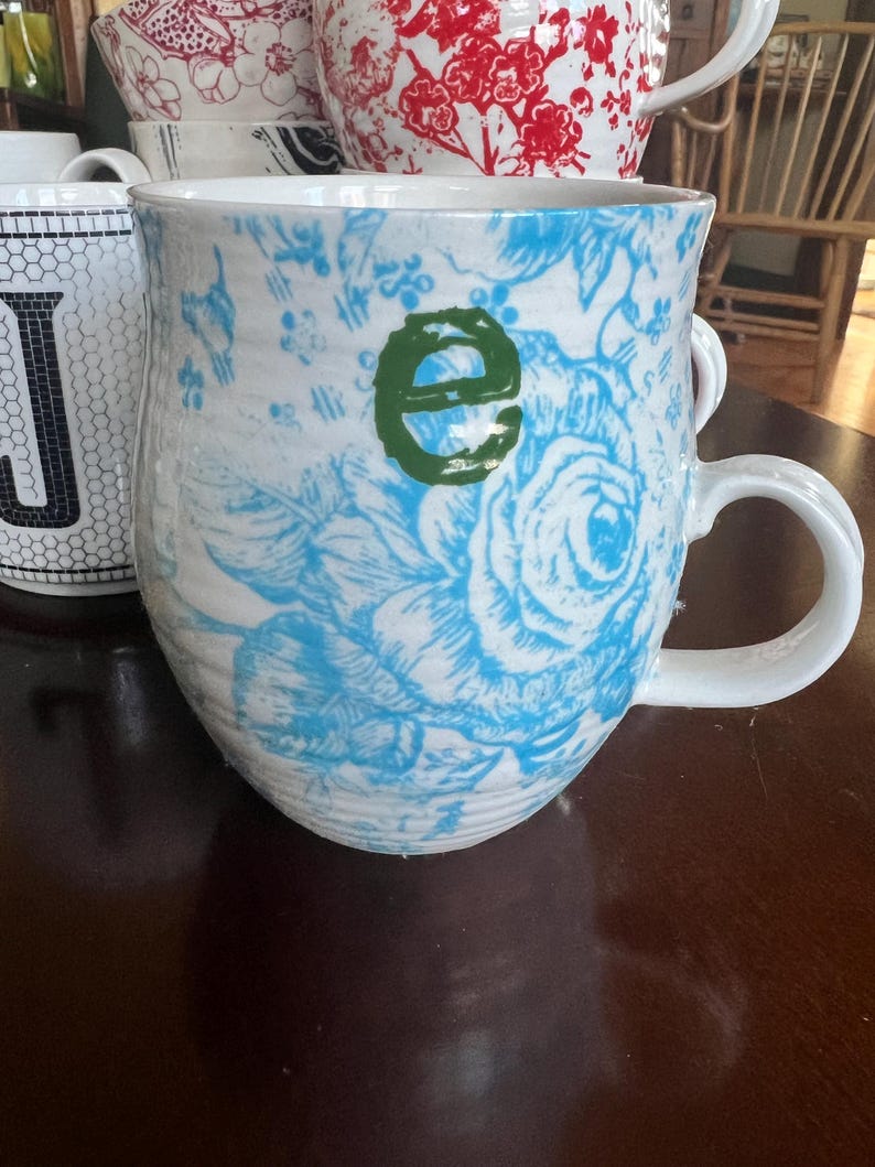 K&ouml;nnte beinhalten: Wei&szlig;er Keramikbecher mit blauem Blumenmuster und einem gr&uuml;nen Buchstaben "e". Der Becher hat einen gro&szlig;en Henkel und geh&ouml;rt zu einer Sammlung von Bechern mit verschiedenen Mustern und Farben. Andere Becher im Hintergrund haben rote Blumenmuster und schwarz-wei&szlig;e geometrische Designs.