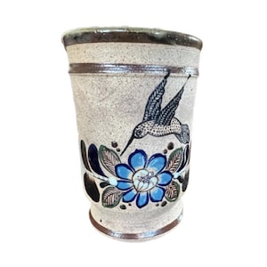 May include: A handcrafted ceramic tumbler with a speckled beige finish. It features a blue floral design with a hummingbird illustration. The rim and base are accented with a dark brown glaze, adding a rustic touch to the piece.