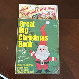 以下が含まれることがあります： 緑色の表紙のヴィンテージ本「Great Big Christmas Book」には、赤いスーツを着たサンタクロースのイラスト、黄色の文字、白いひげが描かれています。その背後には、人形とクリスマスツリーのグラフィックが描かれた「Have a Natural Christmas 84」という小冊子があります。