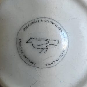 K&ouml;nnte beinhalten: Nahaufnahme des Bodens einer wei&szlig;en Keramiktasse mit blauem Rand. Der Boden zeigt ein kreisf&ouml;rmiges Design mit einer Vogelillustration und dem Text "MICROWAVE & DISHWASHER SAFE", "FABRIQUE EN CHINE" und "MADE IN CHINA".