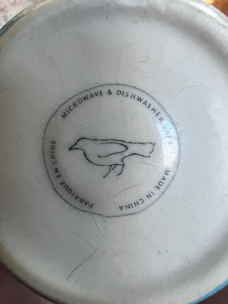 K&ouml;nnte beinhalten: Nahaufnahme des Bodens einer wei&szlig;en Keramiktasse mit blauem Rand. Der Boden zeigt ein kreisf&ouml;rmiges Design mit einer Vogelillustration und dem Text "MICROWAVE & DISHWASHER SAFE", "FABRIQUE EN CHINE" und "MADE IN CHINA".