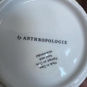 K&ouml;nnte beinhalten: Wei&szlig;er Keramik-Tassenboden mit dem Text "by ANTHROPOLOGIE". Der Boden der Tasse enth&auml;lt auch einen Aufkleber mit dem Text "Made in China", "Fabriqu&eacute; en Chine", "hand wash recommended".