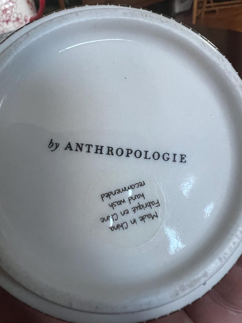 K&ouml;nnte beinhalten: Wei&szlig;er Keramik-Tassenboden mit dem Text "by ANTHROPOLOGIE". Der Boden der Tasse enth&auml;lt auch einen Aufkleber mit dem Text "Made in China", "Fabriqu&eacute; en Chine", "hand wash recommended".