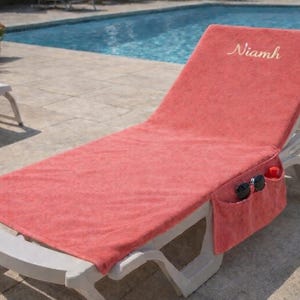 Puede incluir: Funda de tumbona de color coral con el nombre "Niamh" bordado en el reposacabezas. La funda tiene un bolsillo con gafas de sol y una botella. La silla está cerca de una piscina.