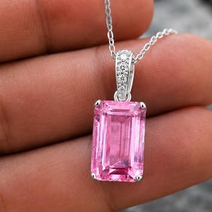 May include: A silver pendant necklace featuring a large, rectangular, emerald-cut pink gemstone. The silver bail is accented with four small, clear, round-cut stones. The pendant hangs from a delicate silver chain with a polished metallic finish.