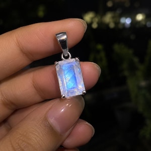 May include: A rectangular moonstone pendant set in a silver-toned metal prong setting. The gemstone displays a translucent, milky white appearance with a distinct, glowing blue adularescence across its surface, attached to a silver-toned bail.