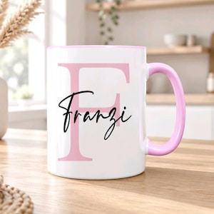 Könnte beinhalten: Eine weiße Keramiktasse mit einem großen, hellrosa Buchstaben F in der Mitte, überlagert von dem Namen Franzi in schwarzer Schreibschrift. Die Tasse hat einen hellrosa Henkel und einen passenden hellrosa Rand für ein klares Design.
