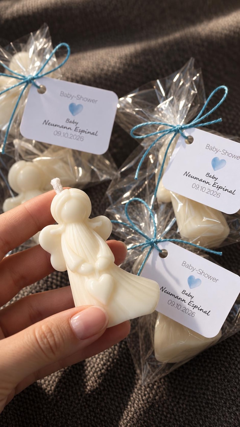 K&ouml;nnte beinhalten: Eine wei&szlig;e engelf&ouml;rmige Kerze mit einem kleinen Herz in den H&auml;nden. Jede Kerze ist einzeln in Klarsichtfolie verpackt, mit blauem Garn verschn&uuml;rt und mit einem wei&szlig;en Etikett versehen, das Baby-Shower, ein blaues Herz, Baby Neumann Espinal und 09.10.2026 zeigt.