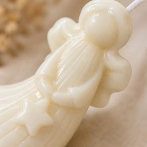 K&ouml;nnte beinhalten: Eine cremefarbene Wachskerze in Form einer stilisierten Figur mit rundem Kopf, flie&szlig;endem Gewand und Fl&uuml;geln. Die Figur h&auml;lt einen f&uuml;nfzackigen Stern in den H&auml;nden, und ein wei&szlig;er Baumwolldocht ragt oben aus dem Kopf der Kerze heraus.
