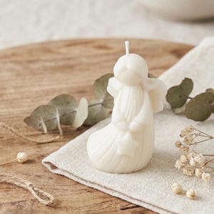 K&ouml;nnte beinhalten: Eine cremefarbene Wachskerze in Form einer stilisierten Engelsfigur mit glattem, rundem Kopf, definierten Fl&uuml;geln und einem langen, flie&szlig;enden Gewand. Die Figur h&auml;lt einen kleinen sternf&ouml;rmigen Gegenstand in den H&auml;nden und ein wei&szlig;er Docht ragt oben heraus.