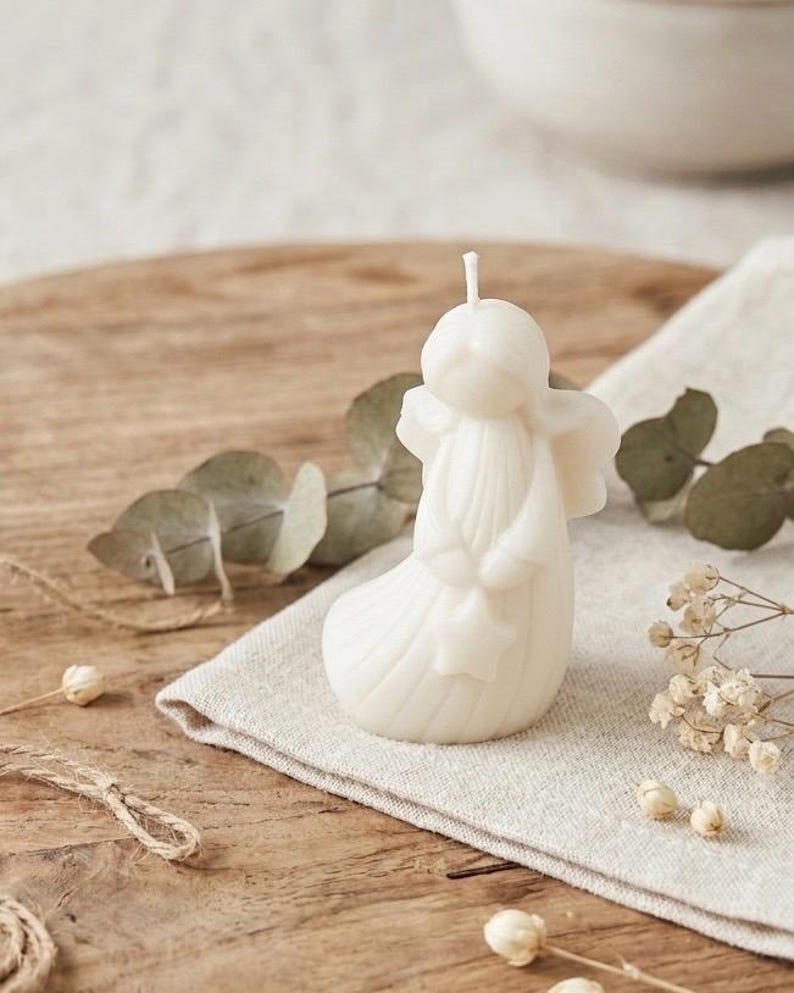 K&ouml;nnte beinhalten: Eine cremefarbene Wachskerze in Form einer stilisierten Engelsfigur mit glattem, rundem Kopf, definierten Fl&uuml;geln und einem langen, flie&szlig;enden Gewand. Die Figur h&auml;lt einen kleinen sternf&ouml;rmigen Gegenstand in den H&auml;nden und ein wei&szlig;er Docht ragt oben heraus.