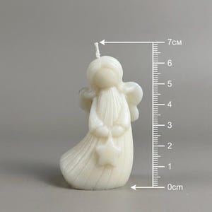 K&ouml;nnte beinhalten: Eine cremewei&szlig;e, engelf&ouml;rmige Kerze mit einem Stern in den H&auml;nden und einem Docht an der Oberseite. Ein vertikales Lineal neben der Kerze zeigt eine H&ouml;he von etwa 7 cm an. Die Kerze hat eine glatte, geformte Oberfl&auml;che mit Fl&uuml;geln und einem flie&szlig;enden Gewand.