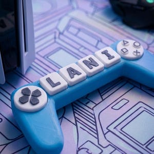 May include: A blue controller-shaped object with the name "LANI" spelled out in white letters. The controller has a D-pad, buttons, and a directional pad. The controller is on a patterned surface with a gaming theme.