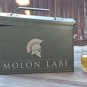 Könnte beinhalten: Eine grüne Metall-Munitionskiste mit einem weißen spartanischen Helm und dem Text "MOLON LABE" auf der Vorderseite. Die Kiste ist geöffnet und steht auf einem Holztisch neben einem Glas mit bernsteinfarbener Flüssigkeit und Eiswürfeln.