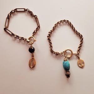 May include: Two gold chain bracelets with charms. The bracelet on the left has a black bead and a gold shell charm. The bracelet on the right has a turquoise bead, a gold alien charm, and a gold bead.