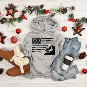 May include: A gray hoodie with a distressed American flag design featuring a silhouette of an off-road vehicle. The hoodie is laid out on a white wooden surface with a pair of brown boots, blue jeans, and a coffee cup.