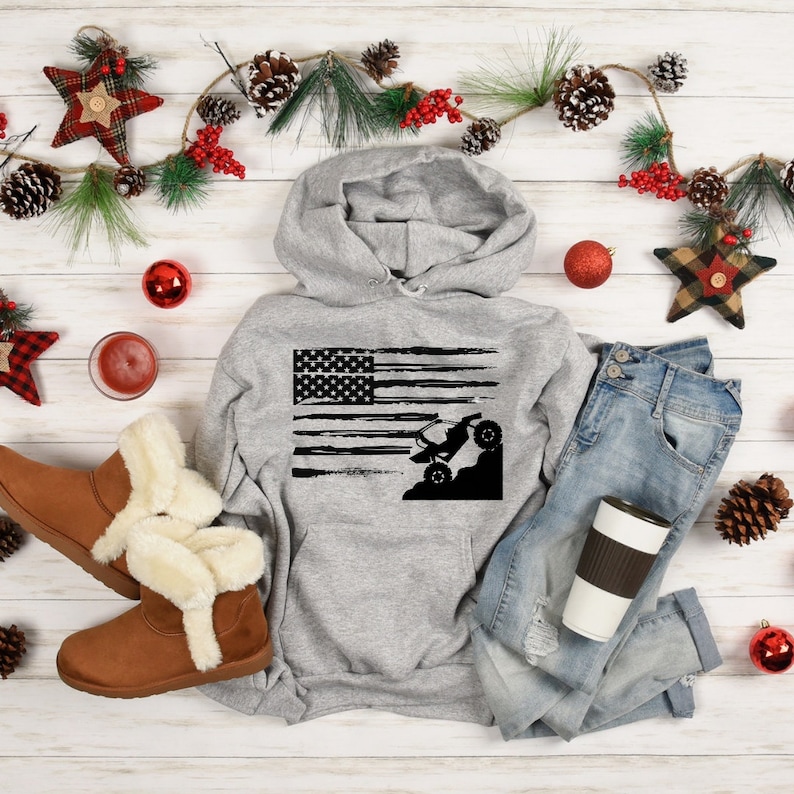 May include: A gray hoodie with a distressed American flag design featuring a silhouette of an off-road vehicle. The hoodie is laid out on a white wooden surface with a pair of brown boots, blue jeans, and a coffee cup.