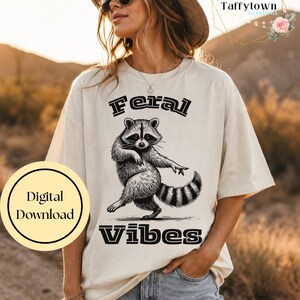 May include: A cream-colored short-sleeve t-shirt featuring a black graphic illustration of a raccoon in a dancing pose. The text "Feral" is printed above the raccoon and "Vibes" is printed below in a bold, stylized black font.