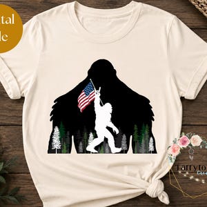 May include: A cream-colored t-shirt featuring a black silhouette of a large creature standing in a forest of green and white trees. The creature holds a small American flag in one hand and makes a hand gesture with the other hand.