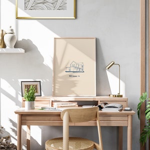 Pu&ograve; includere: Una stampa incorniciata beige chiaro di una casa con un contorno blu. La stampa &egrave; appesa a una parete bianca sopra una scrivania di legno con una sedia. Il testo sulla stampa dice "1004 Summer St."