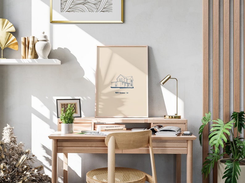 Pu&ograve; includere: Una stampa incorniciata beige chiaro di una casa con un contorno blu. La stampa &egrave; appesa a una parete bianca sopra una scrivania di legno con una sedia. Il testo sulla stampa dice "1004 Summer St."