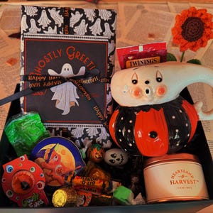 May include: A gift box containing a ghost-patterned wrapped item with a Ghostly Greetings card, a ceramic ghost-shaped mug, a Twinings tea bag, a Heartland Harvest candle, a green candy bag, a round tin, and assorted small Halloween candies.