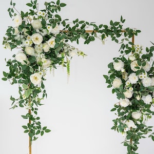 May include: A gold metal rectangular frame arch decorated with two large floral arrangements consisting of white roses, small white daisy-like flowers, and green leafy vines. The flowers are clustered on the top left and right sides.