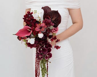 Puede incluir: Un ramo de novia en cascada con calas de color burdeos intenso, anturios de color rojo oscuro, pequeñas orquídeas blancas y amaranto verde colgante. El arreglo incluye varios elementos florales de color rojo oscuro y granate.
