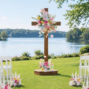 May include: A tall wooden cross standing on a circular base, decorated with a large arrangement of pink, yellow, and white flowers. The cross is positioned in the center of an outdoor aisle lined with white folding chairs and floral bouquets.