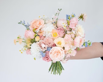 May include: A floral bouquet featuring a mix of peach, soft pink, white, and light blue flowers. The arrangement includes roses, peonies, and small filler blossoms with green stems gathered at the base, creating a rounded, multi-textured display.