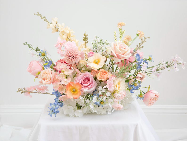 以下が含まれることがあります： パステルカラーの花々が咲き誇る、鮮やかなフラワーアレンジメント。ピンクのバラ、ピーチ色の花、繊細なブルーのアクセントが特徴です。白い表面に花が飾られています。
