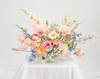 May include: A colourful floral display with a mix of pink, peach, yellow, and blue flowers. The arrangement includes roses, dahlias, and other blooms, creating a soft and romantic look. The flowers are arranged on a white surface.
