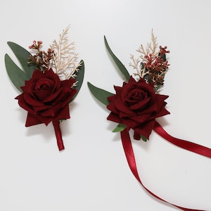 Könnte beinhalten: Zwei bordeauxrote Rosen-Anstecker mit goldenem und grünem Blattwerk. Jeder Anstecker hat ein bordeauxrotes Band.