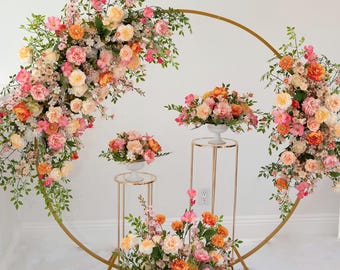 Puede incluir: Un gran marco circular de metal dorado decorado con arreglos de flores rosas, naranjas y crema con follaje verde. Dos soportes de metal dorado de diferentes alturas sostienen centros de mesa florales, con un gran arreglo floral en la base.