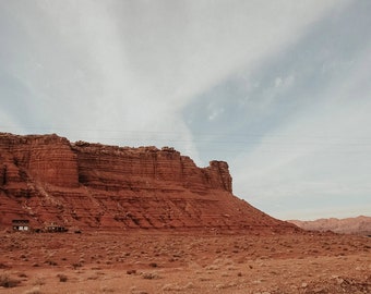 May include: A red rock formation rises up against a cloudy sky. The rock is layered and textured, with a small building at the base. The ground is covered in dry, brown brush.