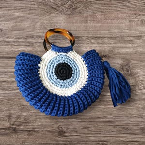 May include: A blue and white crochet handbag with a circular design resembling an eye. The bag features a brown handle and a blue tassel. The bag is made of a thick, textured yarn and sits on a wooden surface.