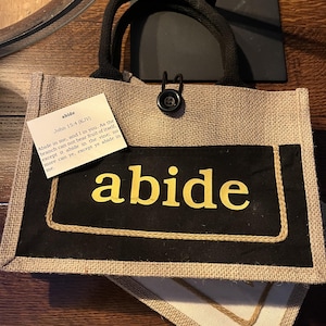 May include: A beige tote bag with black fabric trim and a black button closure. The bag has a quote from the Bible, John 15:4, printed on a white card inside the bag. The quote reads: "Abide in me, and I in you. As the branch cannot bear fruit of itself, except it abide in the vine; no more can ye, except ye abide in me." The word "abide" is printed in large yellow letters on the black fabric trim.