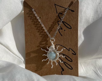 May include: A silver necklace with a wire-wrapped aquamarine stone pendant shaped like a sun. The pendant is on a brown card with the word "Adi" written on it.