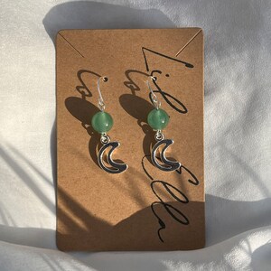 May include: A pair of silver earrings with green stone accents and a crescent moon design.