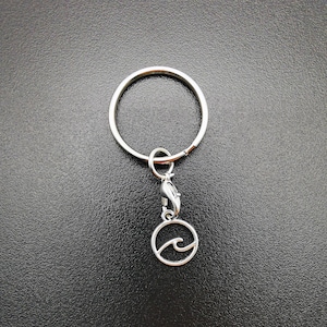 Könnte beinhalten: Ein silberner Schlüsselanhänger mit einem runden Anhänger, der eine Welle zeigt.