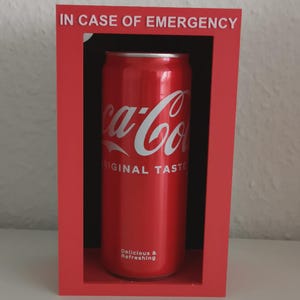 Könnte beinhalten: Eine rote Coca-Cola-Dose mit weißer Schrift, präsentiert in einem roten Rahmen mit dem Text "IN CASE OF EMERGENCY" oben. Auf der Dose stehen die Worte "ORIGINAL TASTE" und "Delicious & Refreshing".