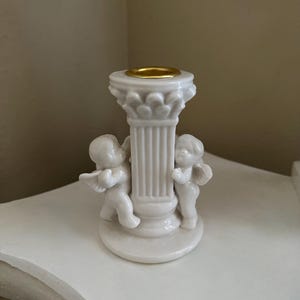 Puede incluir: Un candelabro de cerámica blanca con un diseño de columna clásica y una copa dorada para velas. Dos figuras de querubines alados flanquean la columna. El candelabro es un artículo decorativo.