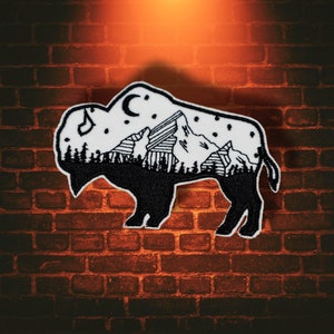 May include: Embroidered patch in the shape of a bison, featuring a black and white mountain and forest scene. The patch includes a crescent moon, stars, and musical notes. The bison is outlined in black.