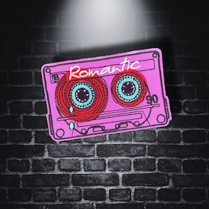May include: A pink embroidered cassette tape patch with red and teal detailing. The word "Romantic" is written in white script across the tape. The patch is set against a dark brick wall background.