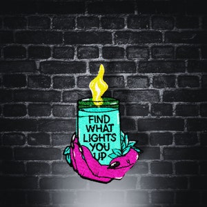 Könnte beinhalten: Eine tealblaue Kerze, die von einer rosa Hand gehalten wird. Die Kerze hat eine gelbe Flamme und den Text "FIND WHAT LIGHTS YOU UP" auf der Vorderseite.